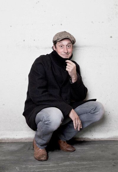 Hans Löw Schauspieler - kniehend vor weißer Wand braune Jacke witziger Blick Mütze Boy Gobert Preis 2004 - 2011 Foto Peter Koehn
